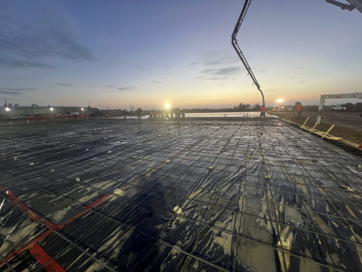 Construction workers prepare a site for Metallic Epoxy Flooring installation at dusk, with machinery and lights in the background.